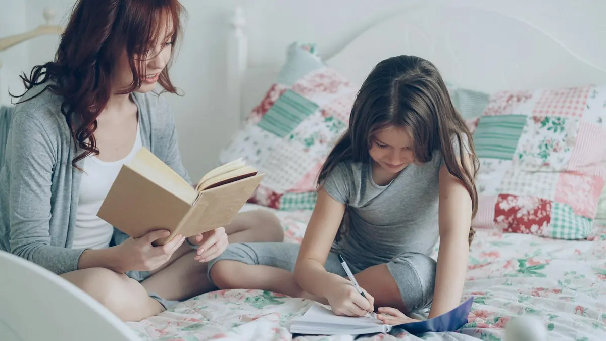 Annesi kitap okurken yanında defterine yazı yazan bir kız çocuğu